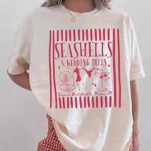 Peut inclure: T-shirt crème avec un motif rouge indiquant "SEASHELLS & WEDDING BELLS" avec des illustrations de coquillage, bikini et cocktail. Le texte "Emma's Bachelorette, Miami, Fl" est également inclus. Le t-shirt est porté avec un short à carreaux rouges et blancs.