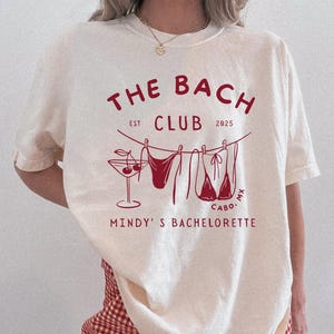 May include: Ivory-colored t-shirt with a dark red graphic design. The design includes the text "THE BACH CLUB EST 2025 CABO MX MINDY'S BACHELORETTE". The graphic also features a martini glass, lingerie, and a clothesline. The shirt is paired with red and white checkered shorts.