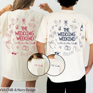 Puede incluir: Dos camisetas de color marfil con un diseño en azul marino y rojo. La parte posterior de las camisetas presenta un círculo de ilustraciones con temática nupcial y el texto "The Wedding Weekend" con los nombres "Mr & Mrs. Smith". La parte delantera de las camisetas tiene el texto "The Smiths" en azul marino.