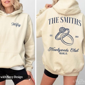 May include: Sand colored hoodie with a navy blue design. The front of the hoodie says "Wifey". The back of the hoodie says "THE SMITHS" with a ring design and "Newlyweds Club MIAMI, FL" below.