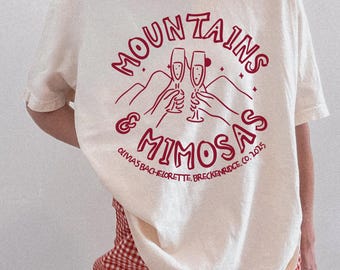 May include: Ivory-colored t-shirt with a dark red graphic that reads "Mountains & Mimosas" with champagne flutes and the text "Olivia's Bachelorette, Breckenridge, CO. 2025". The shirt is paired with red and white checkered shorts.