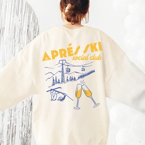 May include: A cream colored sweatshirt with a graphic print on the back. The graphic features a mountain scene with a ski lift, skiers, and champagne glasses. The text "APR&Egrave;S SKI social club" is printed in yellow.