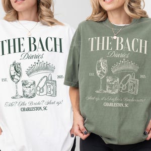 May include: Two white and green t-shirts with the text "The Bach Diaries" and a graphic of a crown, a cocktail glass, a cassette tape, and a glass of ice. The text "EST. 2025" is also on the shirts. One shirt says "Me? The Bride? Shut up! CHARLESTON, SC" and the other says "Shut up, it's Taylor's Bachelorette! CHARLESTON, SC".