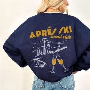 May include: A navy blue sweatshirt with a white graphic of a ski scene and the text "APR&Eacute;S SKI social club" in gold lettering.