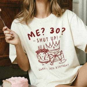 May include: Cream-colored t-shirt with the text "ME? 30? SHUT UP!" and a cake graphic. The shirt is part of a birthday theme, with the words "Gabby 30th Birthday" printed below the graphic. A piece of pink cake sits on a glass cake stand.