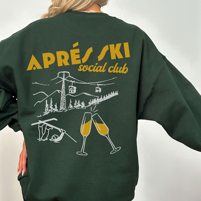 May include: A dark green sweatshirt with a white graphic design on the back. The graphic features a ski lift, mountains, trees, and two champagne glasses. The text "APR&Egrave;S SKI social club" is printed in yellow.