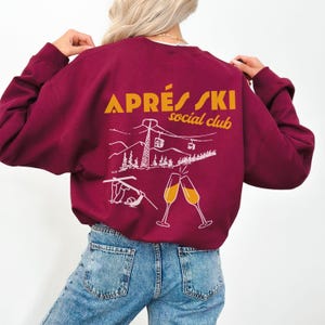 May include: A maroon sweatshirt with a white graphic of a ski scene and the text "APR&Eacute;S SKI social club" in orange.