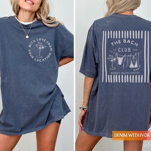 Puede incluir: Una camiseta azul denim con un gr&aacute;fico en la parte delantera que dice "With Love From Your Location" y una copa de c&oacute;ctel. La parte trasera de la camisa tiene un dise&ntilde;o que dice "The Bach Club" con un bikini y una copa de c&oacute;ctel.