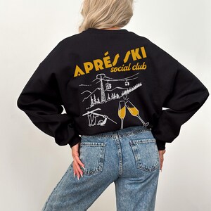 May include: Black sweatshirt with a white graphic of a ski lift, mountains, and champagne glasses. The text "APR&Egrave;S SKI social club" is printed in yellow on the back of the sweatshirt.