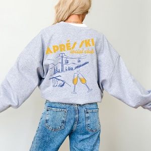 May include: Gray sweatshirt with a graphic print of a ski lift, mountains, and two champagne glasses. The text "APR&Eacute;S/SKI social club" is printed in yellow on the sweatshirt.