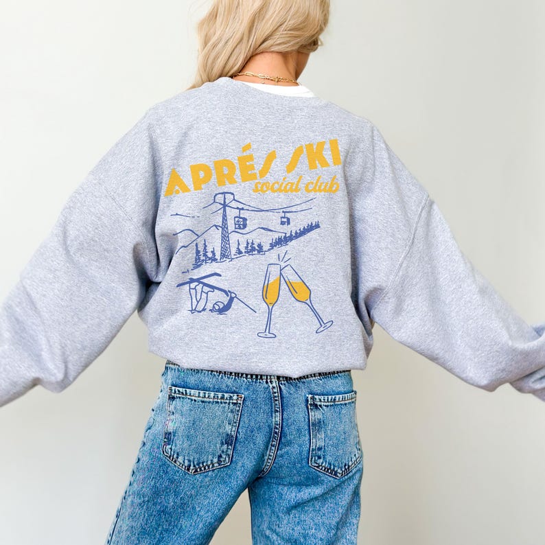 May include: Gray sweatshirt with a graphic print of a ski lift, mountains, and two champagne glasses. The text "APR&Eacute;S/SKI social club" is printed in yellow on the sweatshirt.
