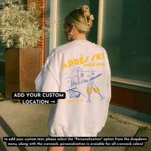 May include: White sweatshirt with a graphic of a ski lift and mountains. The text "APR&Eacute;S/SKI social club" is printed in yellow on the sweatshirt. The text "ADD YOUR CUSTOM LOCATION &rarr;" is printed in black on the bottom left corner of the sweatshirt.