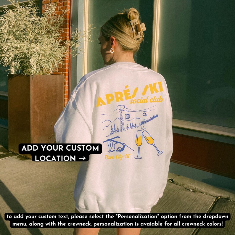 May include: White sweatshirt with a graphic of a ski lift and mountains. The text "APR&Eacute;S/SKI social club" is printed in yellow on the sweatshirt. The text "ADD YOUR CUSTOM LOCATION &rarr;" is printed in black on the bottom left corner of the sweatshirt.