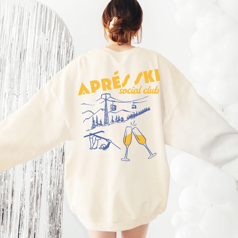 May include: A cream colored sweatshirt with a graphic print on the back. The graphic features a mountain scene with a ski lift, skiers, and champagne glasses. The text "APR&Egrave;S SKI social club" is printed in yellow.