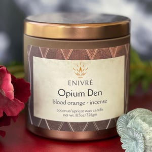 May include: A brown metal candle with a copper-colored lid. The label reads "ENIVRÉ Opium Den blood orange - incense" and "net wt. 11.5oz/326gm". The candle is a coconut/apricot wax blend.