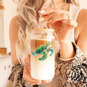 May include: A clear glass tumbler with a bamboo lid and a green sea turtle design. The tumbler contains a layered drink with a straw. The design is a watercolor style, and the tumbler is held by a person.