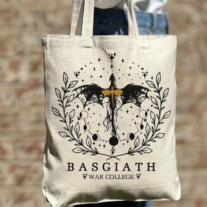May include: A beige tote bag with a black and gold design featuring a dragon, stars, and moons. The text "BASGIATH WAR COLLEGE" is printed on the bag.