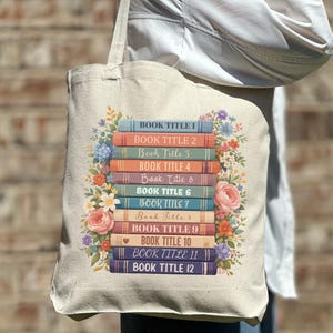 Op de afbeelding: Een beige draagtas met een ontwerp van een stapel boeken omringd door bloemen. Elke boekenrug toont een titel, van 1 tot 12. De tas is gemaakt van een stevige stof, geschikt voor het dragen van boeken en andere items.