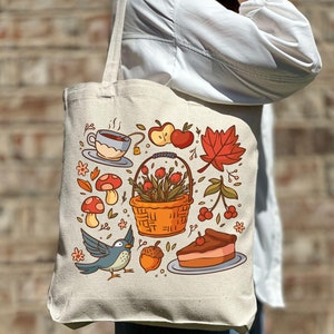 Może przedstawiać: Biała torba z płótna z kolorowym wzorem o tematyce jesiennej. Wzór przedstawia ptaka, grzyby, kosz jagód, kawałek ciasta, filiżankę herbaty i czerwoną liść klonu.