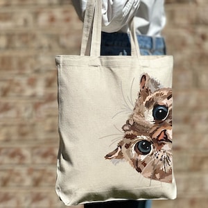 May include: A beige canvas tote bag with a brown and white cat peeking out from the side. The cat has blue eyes and a brown nose.