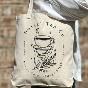 May include: A white canvas tote bag with black text and a graphic design. The text reads "Suriel Tea Co. Hot or Cold, Always Fresh. High Lord Hot Tea". The graphic design features a hand holding a cup of tea with a crescent moon and stars in the background.