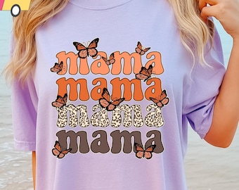 May include: A lavender t-shirt with the text "mama mama mama mama" in a retro font with butterfly graphics. The butterflies are orange, brown, and white.