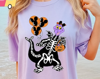 May include: Purple dragon skeleton holding a pumpkin bucket and balloons. The dragon is wearing a purple hat. The design is on a lavender t-shirt.