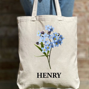May include: A white canvas tote bag featuring a blue forget-me-not flower design and the name "HENRY" printed in black.