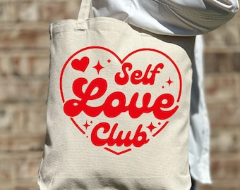 May include: A white canvas tote bag with red text that reads "Self Love Club" inside a heart shape with stars.