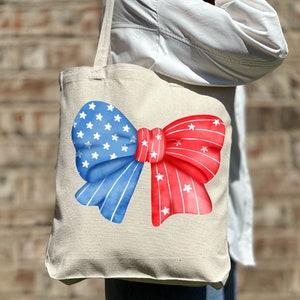 May include: A white canvas tote bag with a red, white, and blue bow design. The bow is made of fabric with stars and stripes.