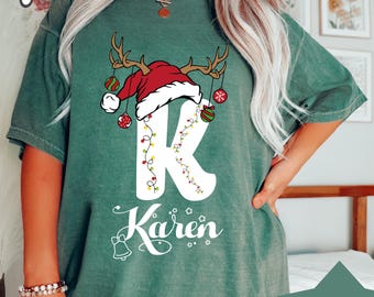 Pode incluir: Uma camiseta azul abeto com um design de Natal. O design apresenta a letra "K" branca decorada com um gorro de Papai Noel, chifres e enfeites. O nome "Karen" está escrito abaixo. A camiseta tem mangas curtas.
