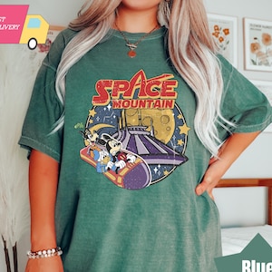 Puede incluir: Una camiseta verde con un gráfico de estilo vintage de Mickey Mouse y Pluto montando Space Mountain. El texto "Space Mountain" está impreso en blanco en la camiseta.