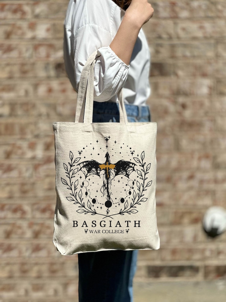 Op de afbeelding: Een beige tote bag met een zwart en gouden ontwerp met een draak, sterren en manen. De tekst "BASGIATH WAR COLLEGE" is op de tas gedrukt.