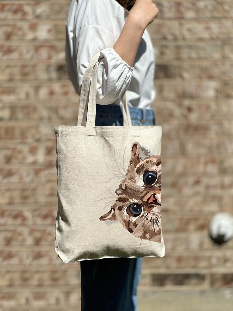 May include: A beige canvas tote bag with a brown and white cat peeking out from the side. The cat has blue eyes and a brown nose.