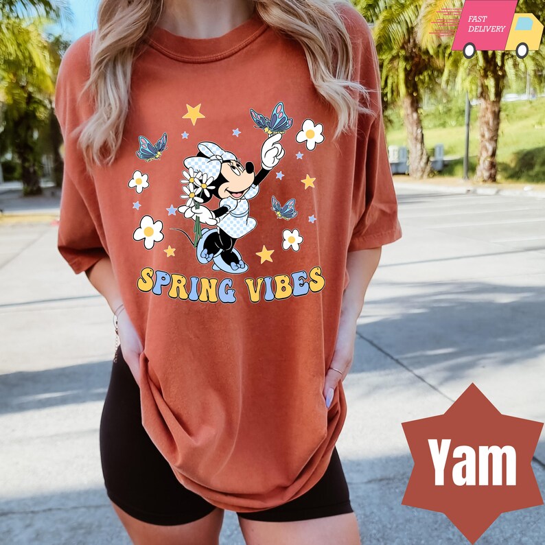 Pode incluir: Uma camiseta cor ferrugem com uma imagem de desenho animado do Mickey Mouse segurando uma flor e borboletas. O texto "SPRING VIBES" &eacute; impresso em amarelo na camiseta.