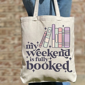 May include: A white canvas tote bag with the text "my weekend is fully booked" in dark blue. The text is surrounded by stars and a stack of colorful books.