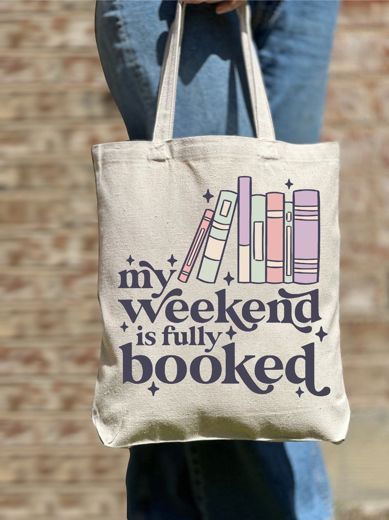 May include: A white canvas tote bag with the text "my weekend is fully booked" in dark blue. The text is surrounded by stars and a stack of colorful books.
