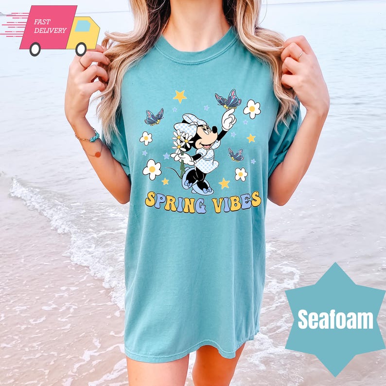 Pode incluir: Uma camiseta azul turquesa com um gr&aacute;fico de Minnie Mouse segurando uma flor e borboletas. O texto "Spring Vibes" est&aacute; impresso abaixo do gr&aacute;fico.