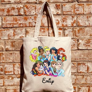 Pu&ograve; includere: Una borsa tote in tela beige con una stampa colorata di principesse Disney. Le principesse sono disposte in cerchio con i loro nomi scritti sotto. La borsa ha un nome personalizzato "Emily" stampato in basso.