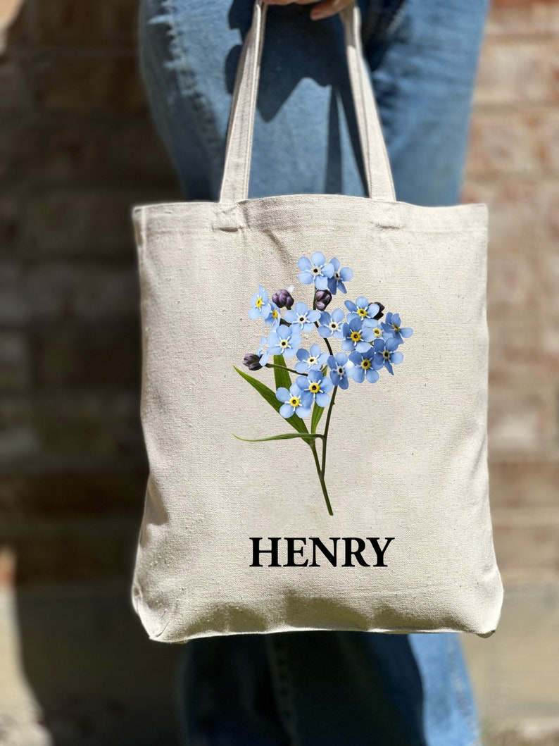May include: A white canvas tote bag featuring a blue forget-me-not flower design and the name "HENRY" printed in black.