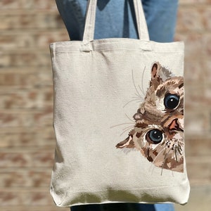 May include: A white canvas tote bag with a brown and white cat peeking out from the right side. The cat has blue eyes and a pink nose.