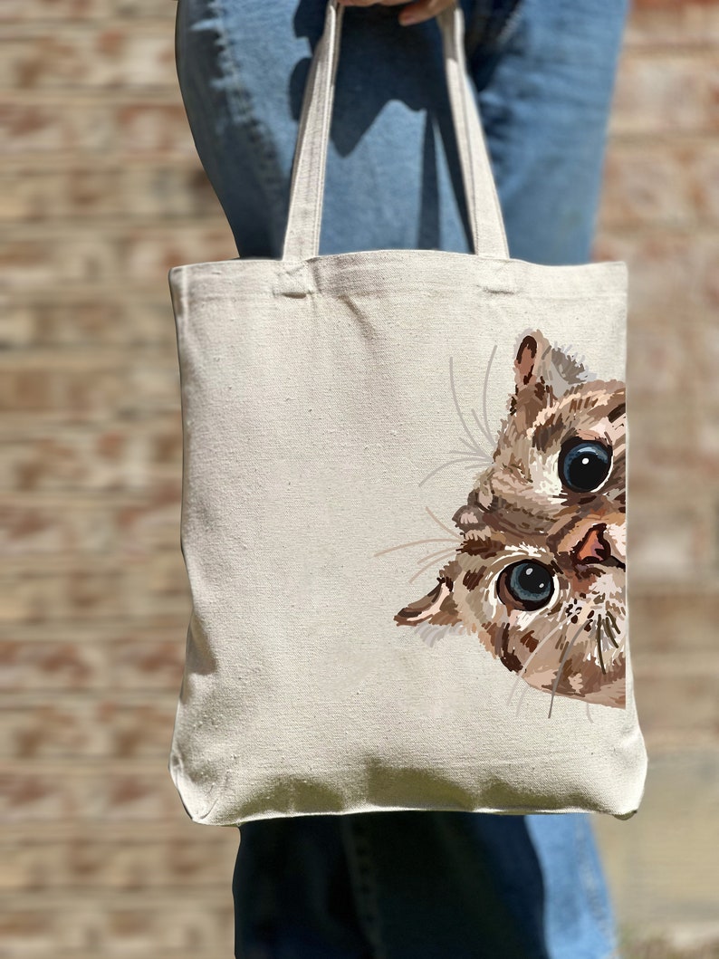 May include: A white canvas tote bag with a brown and white cat peeking out from the right side. The cat has blue eyes and a pink nose.