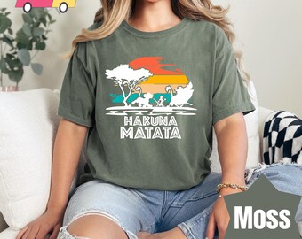 May include: Moss green t-shirt with a colorful graphic featuring a sunset, trees, and animal silhouettes. The text "HAKUNA MATATA" is printed in white below the graphic. The shirt is made of a soft material.