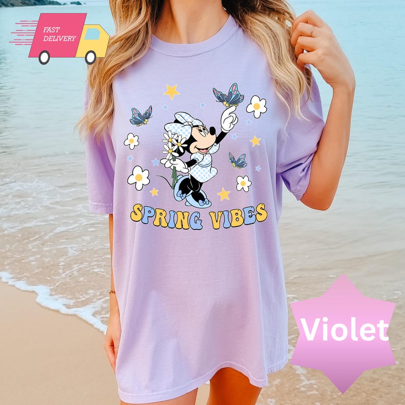 Pode incluir: Uma camiseta roxa com um gr&aacute;fico de Minnie Mouse segurando flores e borboletas. O texto "Spring Vibes" &eacute; impresso abaixo do gr&aacute;fico.