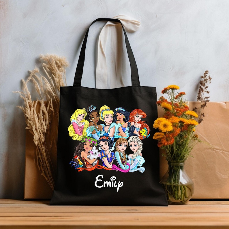 Pu&ograve; includere: Borsa tote nera con un'illustrazione colorata di principesse Disney, tra cui Cenerentola, Ariel e Moana. La borsa ha un manico bianco e il nome "Emily" &egrave; stampato in bianco in basso.