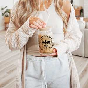 Op de afbeelding: Een doorzichtige glazen beker met een bamboe deksel en een rietje. De beker heeft een zwart-gouden afbeelding met de tekst "BASGIATH WAR COLLEGE DRAGON RIDER". Het glas is gevuld met een lichtbruine vloeistof.