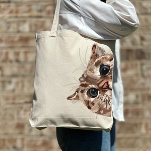 May include: A beige canvas tote bag with a brown and white cat peeking out from the side. The cat has blue eyes and a curious expression.