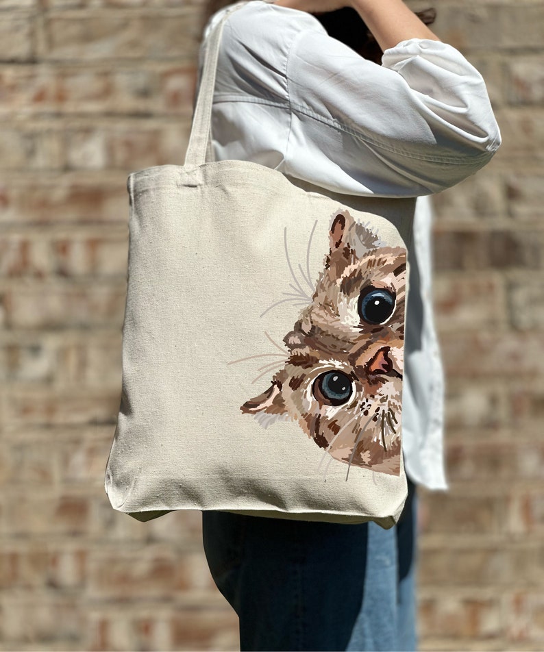 May include: A beige canvas tote bag with a brown and white cat peeking out from the side. The cat has blue eyes and a curious expression.