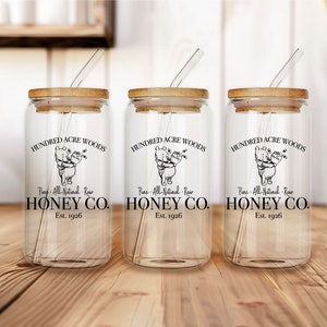 May include: Three clear glass jars with bamboo lids and straws. Each jar has a black and white illustration of Winnie the Pooh and Piglet with the text "Hundred Acre Woods", "Pure All-Natural Raw Honey Co.", and "Est. 1926".