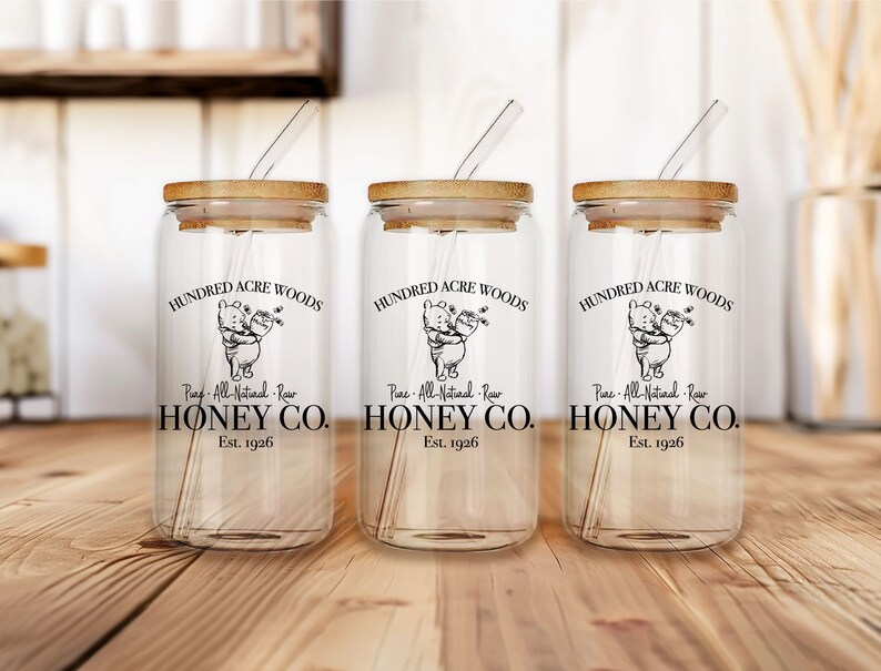 May include: Three clear glass jars with bamboo lids and straws. Each jar has a black and white illustration of Winnie the Pooh and Piglet with the text "Hundred Acre Woods", "Pure All-Natural Raw Honey Co.", and "Est. 1926".